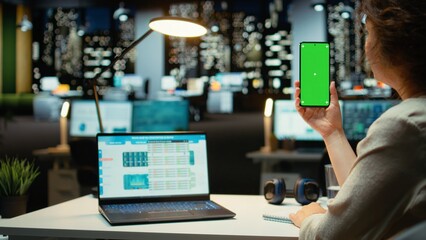 Businesswoman holds phone with chroma key at her desk for task management late at night, business administration alone in the office. Analyzing and planning budgeting reports. Camera A.