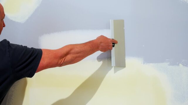 Construction worker spreading joint compound on the drywall wall using a metal taping knife. Wall is smoothed and finished as part of a home renovation.