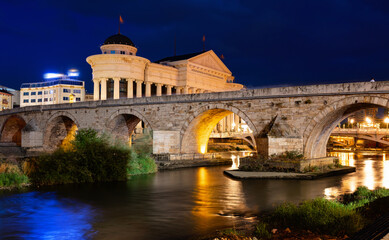 Obraz premium Historic Stone Bridge in Skopje with glowing street lamps reflecting in Vardar River on summer evening, surrounded by illuminated architecture