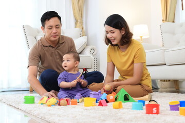 An asian family of three, a woman and two men, are playing with colorful toys on the floor. Asian baby boy is holding a toy and the parents are clapping. Scene is happy and playful