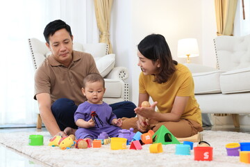 Fototapeta premium An asian family of three, a woman and two men, are playing with colorful toys on the floor. Asian baby boy is holding a toy and the parents are clapping. Scene is happy and playful