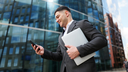 Businessman using smartphone while holding laptop outside modern office building