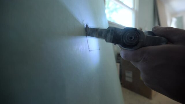 Close-up shot of a construction worker using an oscillating multi-tool to cut a rectangular opening in drywall. Dust particles are visible as the blade slices through.