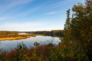 A wide, calm, blue river with colorful fall foliage on both sides. The trees are various shades of red, gold, yellow, and orange. The treeline is to the sandy riverbanks. 