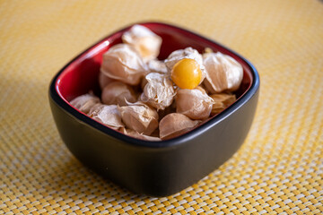 A red and black square porcelain bowl filled with ground cherries, goldenberries, or Cape Gooseberries on a yellow cloth. The cherries are called poha and are edible. The outside is a dry papery husk.