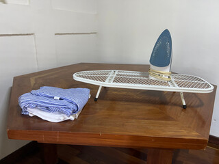 An iron placed on a metal ironing board which is placed on another wooden table; there is also clothing, ready to be ironed.