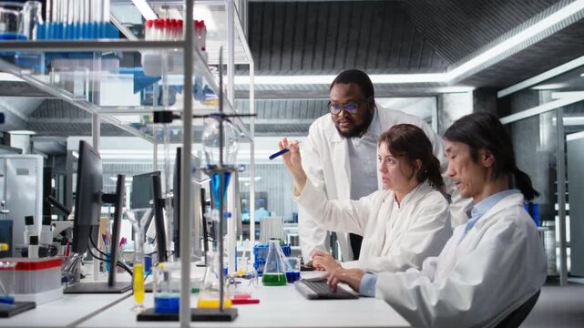 Multiethnic chemical engineers using laboratory glassware filled with indicator solution at workstation. Diverse team of lab specialist picking vial with chemical fluid used for experiments, camera A