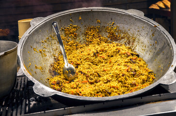 Large metal cauldron filled with a traditional pilaf rice dish. A serving spoon rests in the center of the steaming, savory food with carrots and meat