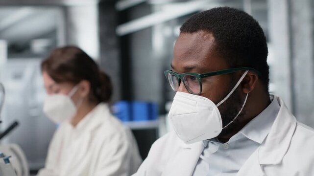 Lab scientist removing protective face mask to scratch beard while using PC. Expert wearing medical mask in contagious workspace studies DNA strand mapping with genomics analysis program, camera B
