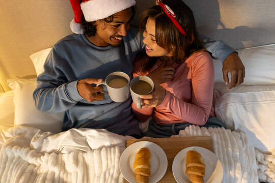 Diverse couple in festive gear sitting on bed toasting mugs over croissants on breakfast tray - Powered by Adobe