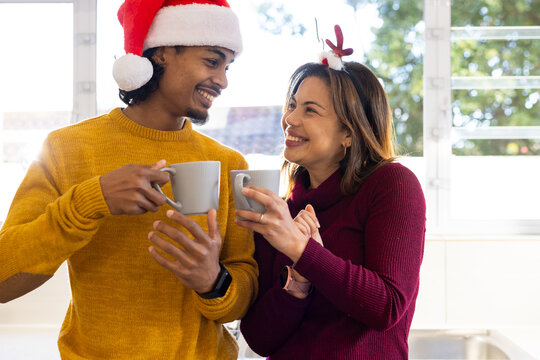 Diverse couple clinking ceramic mugs while wearing Santa hat and antler headband by kitchen window