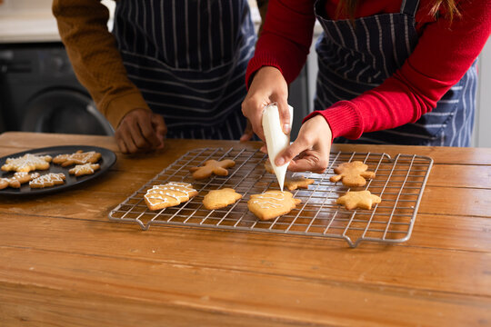 Diverse mother and son decorating gingerbread cookies on rack at kitchen counter using piping bag