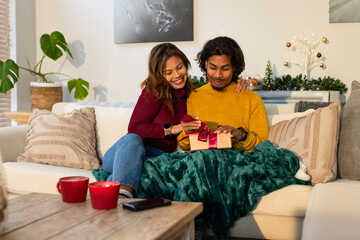 Diverse couple unwrapping ribboned gift box on sofa with throw and red mugs in living room