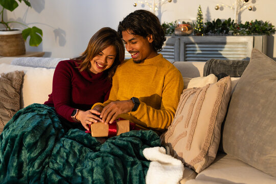 Couple sitting on sofa with blanket opening brown ribboned gift box in festive living room