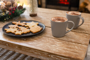 Iced gingerbread cookies resting on plate on wooden table near glowing fireplace, mugs steaming