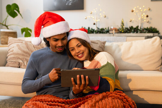 Diverse couple sitting together on plush blanket in living room holding tablet wearing Santa hats