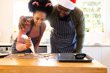 Diverse couple wearing aprons using piping bag decorating gingerbread cookies on kitchen island