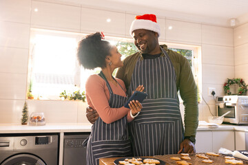 African American couple wearing aprons hugging in kitchen while checking smartphone by cookie tray