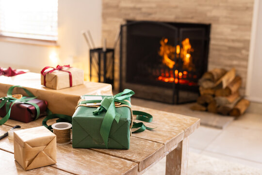 Green-wrapped gift box sitting on table near glowing fireplace, with spools of ribbon and twine