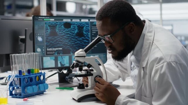 Microbiologist in laboratory inspecting specimen slide using optical device, analyzing on PC. Man reviewing cytology slide for clinical testing procedure, inputting results on computer, camera B