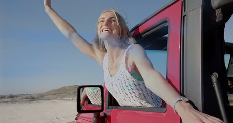 Smiling woman stretching arms out of red offroad vehicle at sand dunes with crochet top, wristwatch