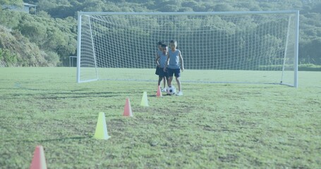 Practicing three boys wearing blue tops kicking soccer ball toward goal on valley pitch, with cones