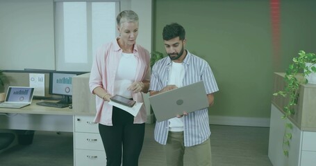 Office colleagues examining charts in office, with monitors, laptop, tablet and printed documents