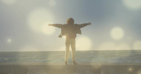 Standing woman stretching arms toward ocean horizon at concrete seawall, with lens flare