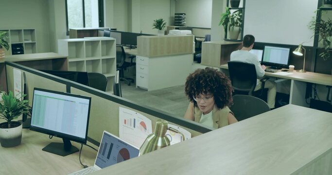 Curly-haired worker in sleeveless top reviewing charts on monitor at desk with lamp, copy space