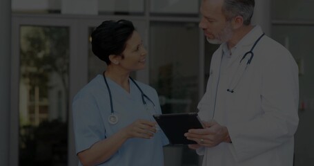 Doctor in lab coat and nurse in blue scrubs discussing at hospital doors, with tablet, stethoscopes