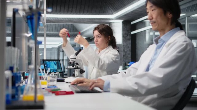 Laboratory employee looks at blood sample and PH indicator in test tubes. Clinical lab researcher analyzing sanguine fluid specimen and blue chemical liquid in vial used for medical research, camera A