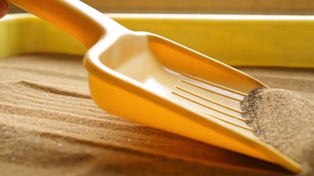 Bright yellow plastic scoop digging into a pile of fine brown sand with a yellow object in the blurry background