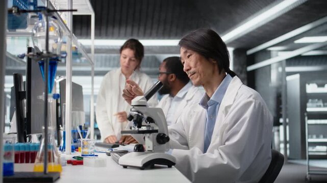 Senior microscopist examining microscope glass slide in laboratory to view cells. Asian research facility employee checks sample slide before imaging process using optical device, camera A