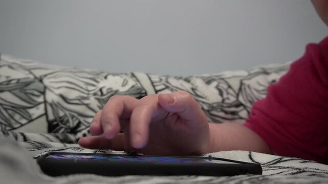 A man is lying on his stomach with a phone in his hands on a gray blanket. Close-up of hands with a black smartphone. Side view.