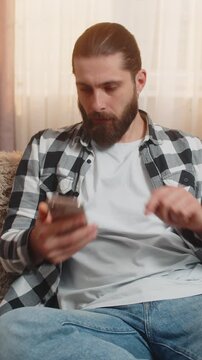 Young man sitting at home, receiving crucial smartphone call quarrel and placing device on knee with visible tension. Caucasian guy gestures irritably, pushing a firm argument through raised voice.