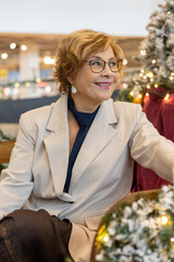 Woman with glasses smiles while sitting in a decorated area during the holiday season in a shopping center