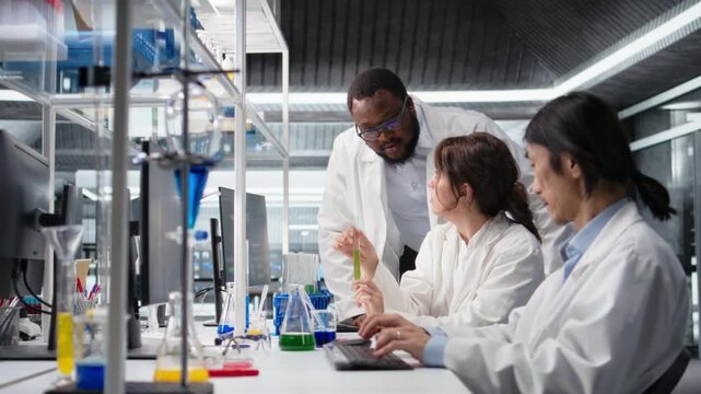 Multiethnic research facility team using colorful chemical solutions in glassware. Diverse teamworking analytical chemists in laboratory looking at reagent vial used for experiment, camera A