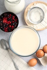 Liquid dough in bowl, berries and ingredients on light grey table, flat lay