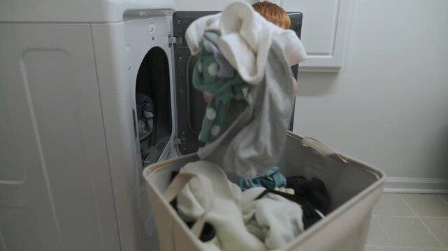 Toddler red hair exploring washing machine laundry basket filled mixed clothing Little boy reaching into front-load washing machine while standing next to a full laundry basket in home setting
