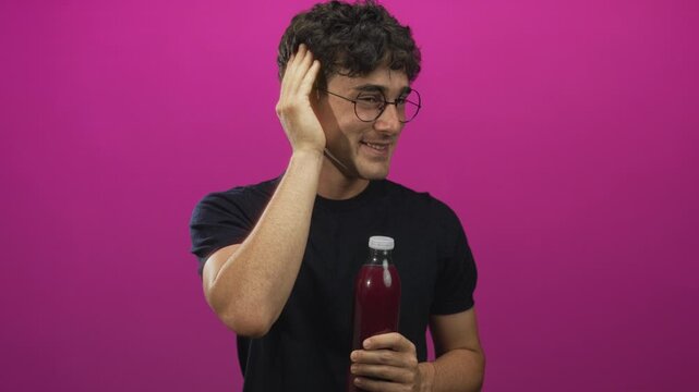 Man wearing glasses and a dark t shirt holding a bottle of juice, hand to ear and smiling in studio; taste test playful.