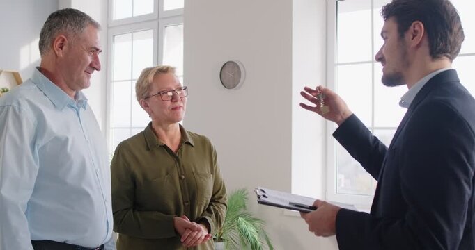 Male real estate agent gives keys to new home owners, happy elderly couple clients, realtor or broker congratulates happy mature spouses with house purchase, tenancy agreement, enjoy ownership moving 