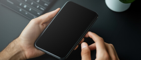 Modern black smartphone held by male hand on dark desk with laptop keyboard and plant in soft light conveying focused mood and minimal workspace