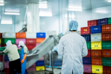 A male factory inspector in a lab coat, hairnet, and safety glasses stands and observes a busy food...