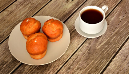 Three vanilla cupcakes in a round plate and a cup of hot tea on a wooden table.