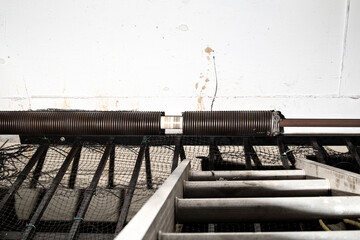 Garage door spring broken on overhead sectional door. Parking gate torsion spring is torn apart close to the ceiling, inside. Emergency clamp and replacement needed. Selective focus.