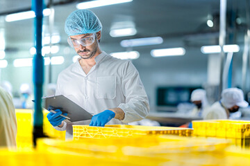 A male factory inspector in a lab coat, hairnet, and safety glasses is checking inventory. He holds...