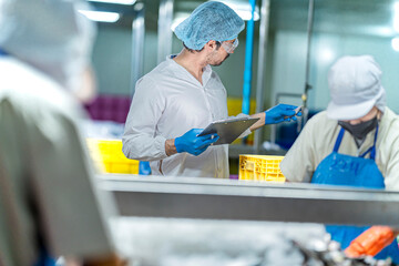 A male inspector in a lab coat, hairnet, and safety glasses checks a small fish. He is holding a clipboard and doing a quality control check in a food processing factory.