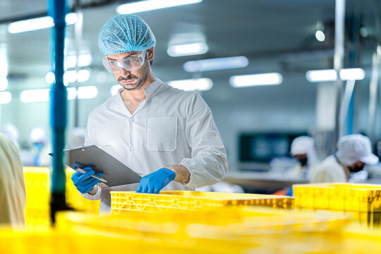 A male factory inspector in a lab coat, hairnet, and safety glasses is checking inventory. He holds a clipboard and is monitoring the production area in a food plant.