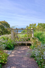 Scenic garden pathway leading to a rustic wooden arbor constructed from natural branches, surrounded by blooming white flowers and lush greenery