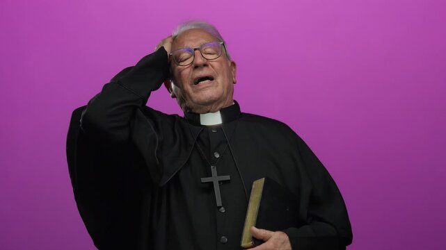 Senior priest in black robe, holding a book, stands over an isolated pink background with a thoughtful expression, highlighting faith and contemplation.